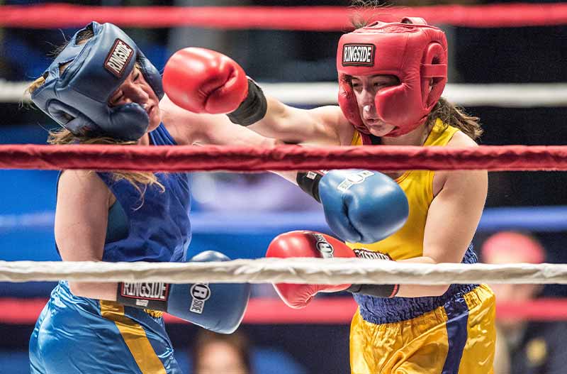 Two young women wearing boxing pads and gloves box in a boxing ring. One woman throws a punch towards the other.