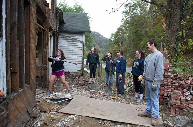One girl wearing protective goggles swings a hammer toward a dilapidated house while five other students stand back and watch.