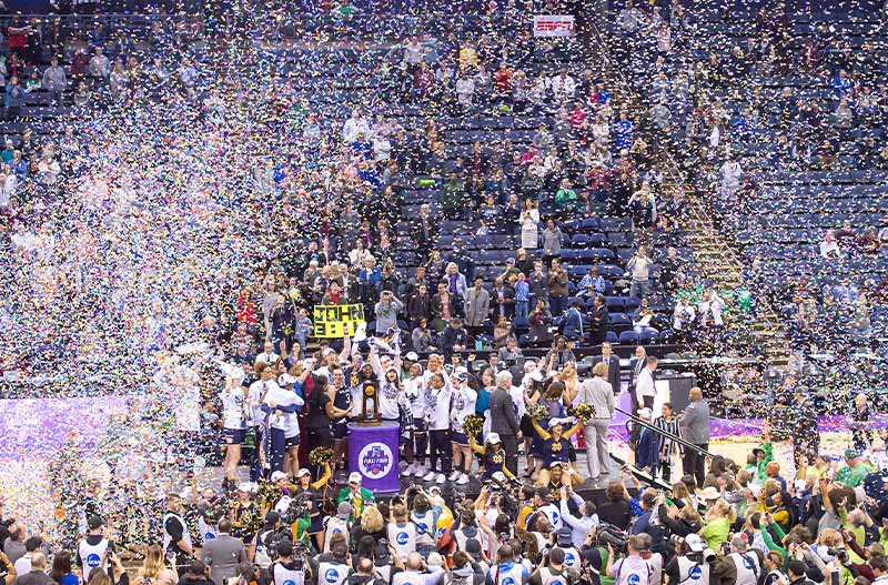 The Notre Dame Women's Basketball team stands in the middle of a basketball quart and celebrates while confetti falls from the ceiling.