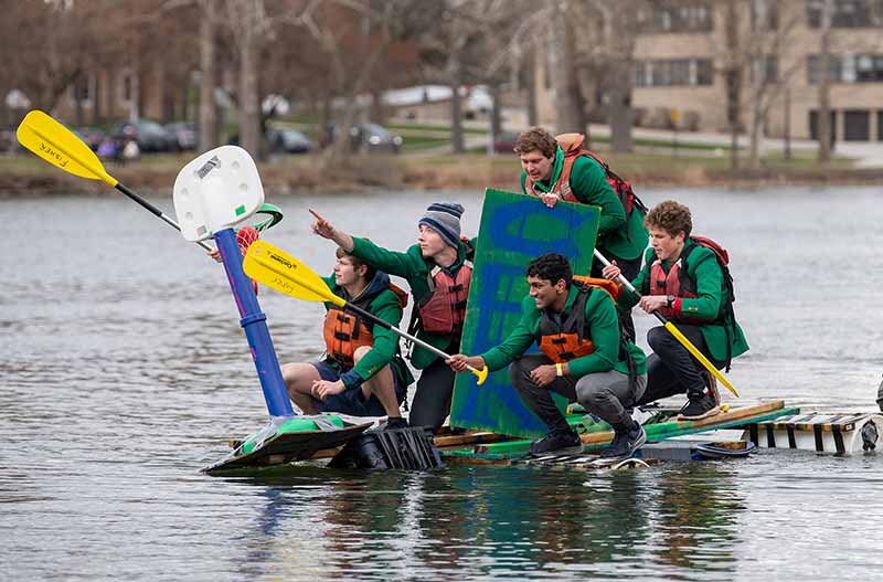 A group of five male students squat on top of a homemade boat. One student rows as two people point onward with their paddles.