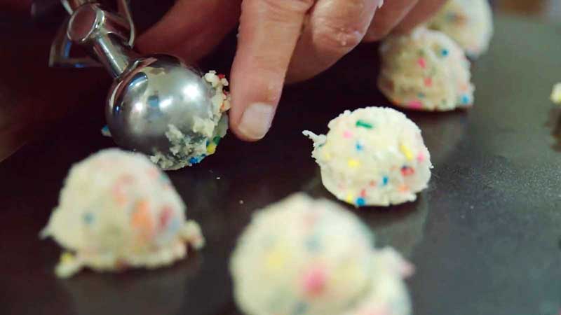 A close-up of hands using an ice cream scooper to place rainbow sprinkle-filled sugar cookie dough balls onto a baking sheet.