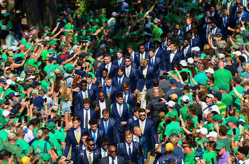 The Notre Dame football team, wearing navy blue suits, walks between large groups of fans taking photos and cheering.