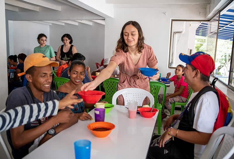 A woman passes out bowls of soup to a table of people.