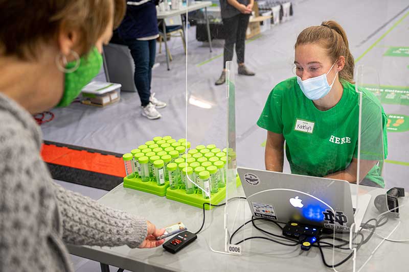 A masked student worker sits behind a plexiglass divider as she assits a staff member who is checking in to do their saliva test.