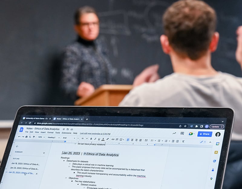 In the foreground a computer screen taking notes while Kirsten Martin interacts with student in the background.