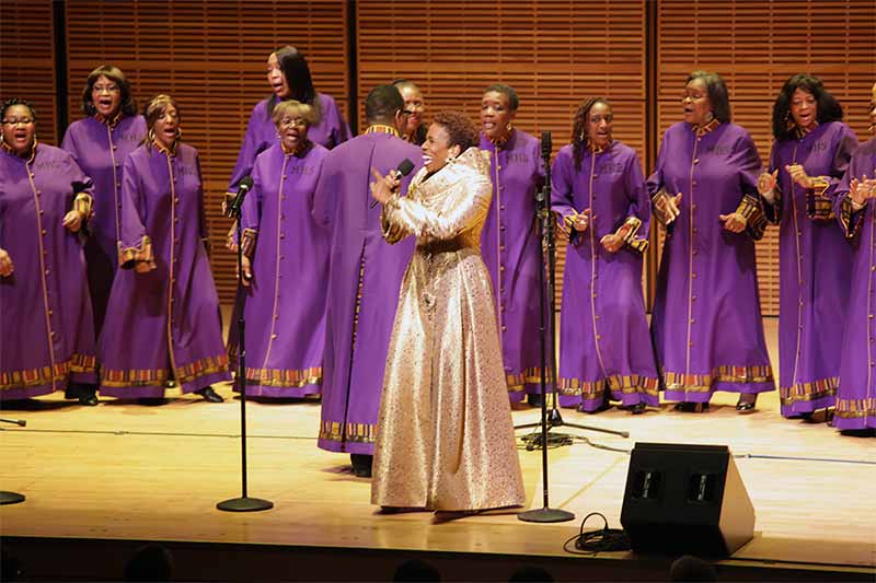 A group of women on stage singing wearing choir robes, one woman's robe is gold.