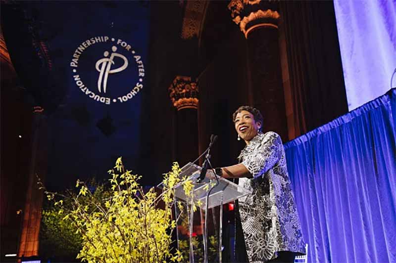 A woman stands at a podium speaking to guests at an award benefit. There are bright, yellow flowers in front of the podium and a blue curtain behind the speaker.