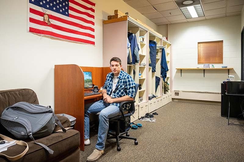 A young man sits at a desk with a computer. A backpack sits on a chair beside him and an American flag hangs above him.