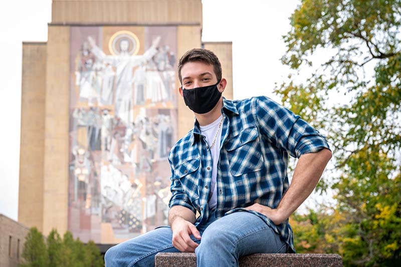 A young masked man sits in front of Touchdown Jesus Hesburgh Library.