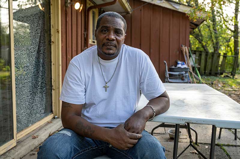 A man wearing a white t-shirt and cross necklace sits at a picnic table and looks at the camera.