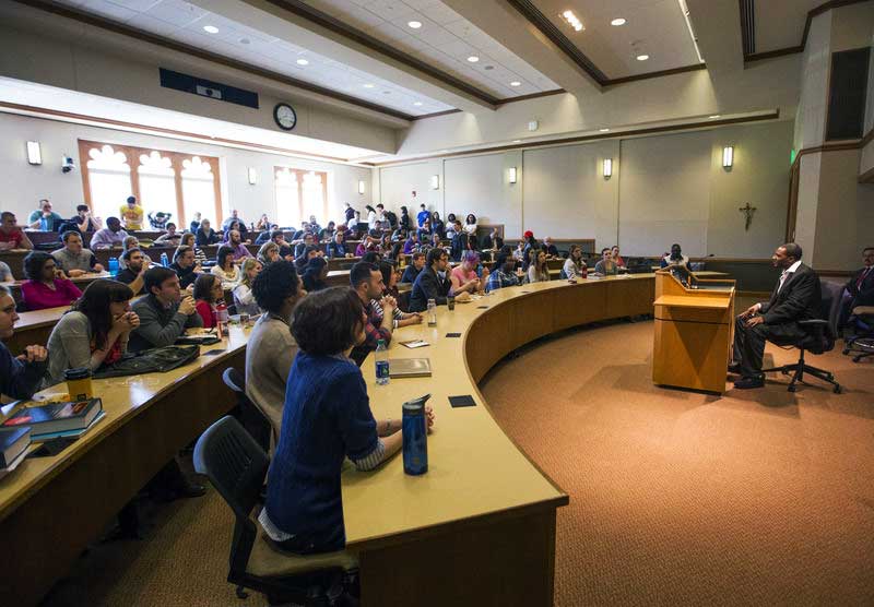 A packed full classroom of students listen to a man tell a story in front of the room.