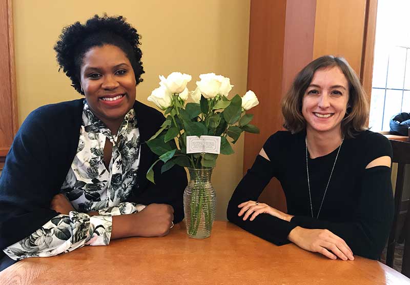 Two women sit a table, a vase of white roses and a note in the middle.