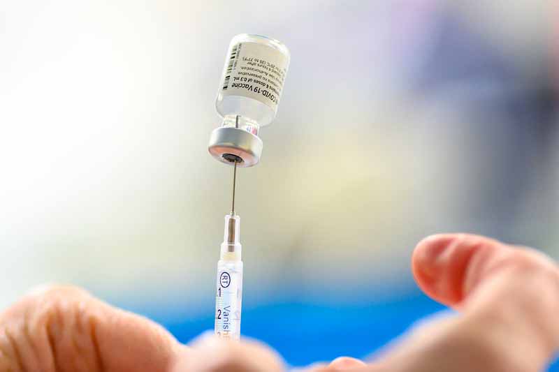 A man, blurred in the background, holds up a vial and needle.