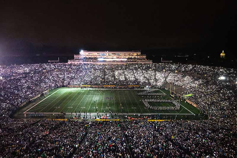 Notre Dame Stadium lit up at night by the fans phone flashlights. In the background, the Golden Dome  glows against the night sky.