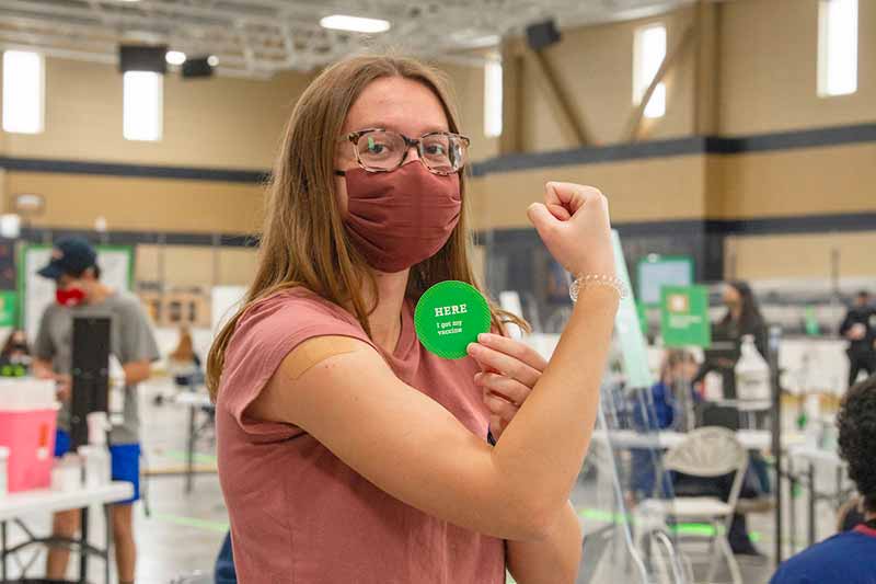 A woman wearing a mask, flexes with a bandaid on her arm, while holding a sticker that says 'Here I got my vaccine'.